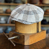 Textured Linen Hatteras flat cap featuring a classic plaid pattern in neutral tones, displayed on a wooden hat block with a blurred background of various hats.