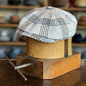 Textured Linen Hatteras flat cap featuring a classic plaid pattern in neutral tones, displayed on a wooden hat block with a blurred background of various hats.