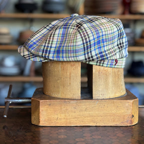 The Brightest Hatteras is a colorful plaid newsboy cap displayed on a wooden hat block in a workshop setting.