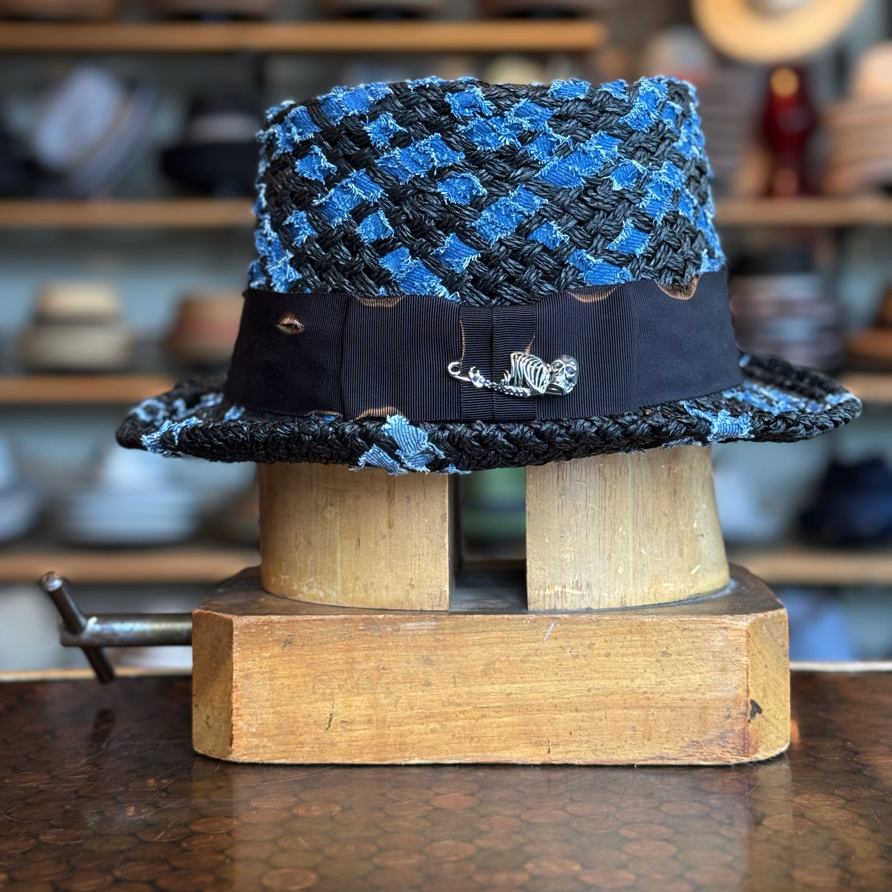 The Denim Top Hat in a woven blue and black pattern with a black band featuring a metallic fish pin, displayed on a wooden hat block in a workshop setting.