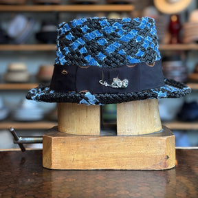 The Denim Top Hat in a woven blue and black pattern with a black band featuring a metallic fish pin, displayed on a wooden hat block in a workshop setting.
