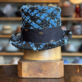 The Denim Top Hat featuring a textured blue and black woven pattern with a wide black band around the crown, displayed on a wooden hat block in a rustic setting.