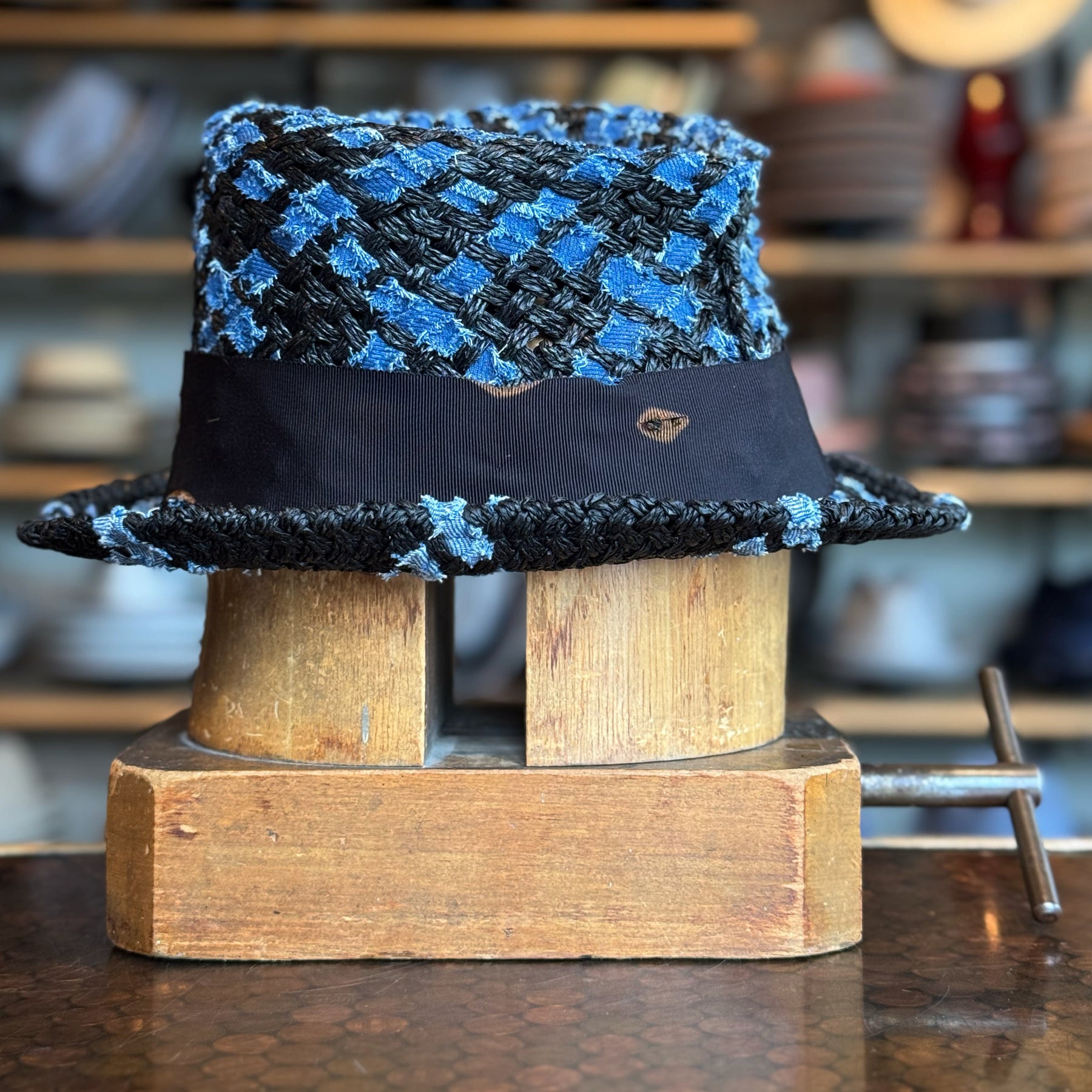 The Denim Top Hat featuring a textured blue and black woven pattern with a black band, displayed on a wooden hat block in a cozy workshop setting.