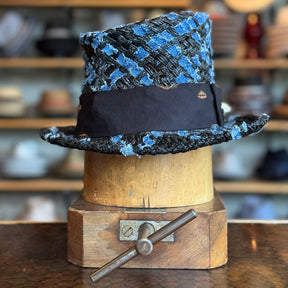 The Denim Top Hat displayed on a wooden hat block, featuring a textured blue and black woven fabric with a black band, styled as a classic top hat accessory.