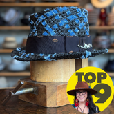 The Denim Top Hat featuring a textured blue and black woven pattern with a black fabric band and a metallic detail, displayed on a wooden hat stand.