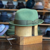 Side of Tino wool felt fedora in kelp green with matching grosgrain band and subtle metal accents, displayed on a wooden hat block in a hat workshop background.