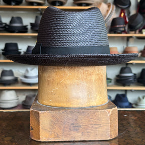 Torino black woven fedora hat with a sleek black band displayed on a wooden hat block in a shop filled with various hats in the background.