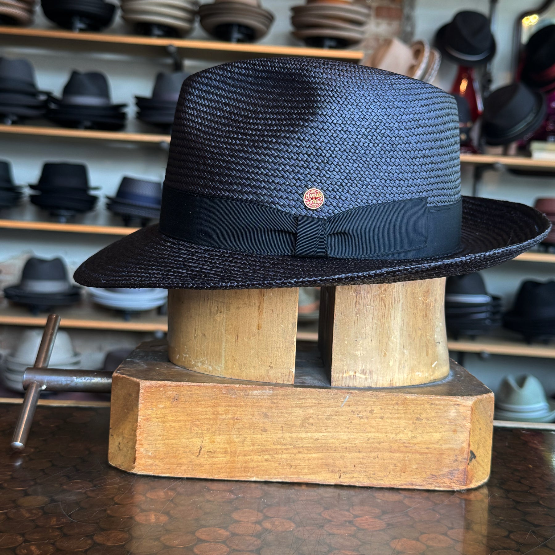 Black woven flat brim hat with black ribbon and a small round emblem, displayed on a wooden hat block in a shop with shelves of various hats in the background, Torino model.