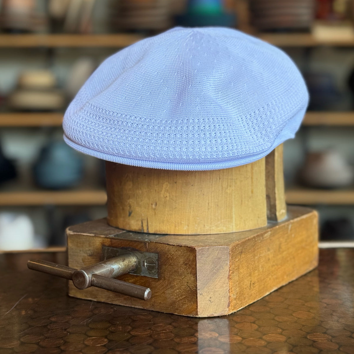 Tropic 504 Ventair white knit flat cap displayed on a wooden hat block with a blurred background of other hats on shelves.
