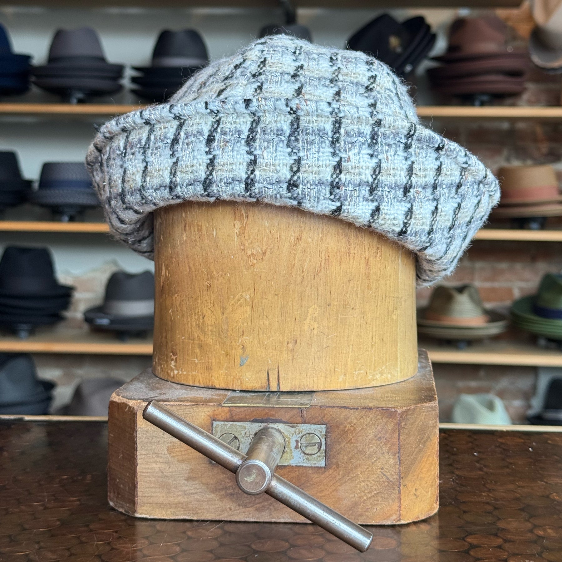 Tweed Sailor Cap displayed on a vintage wooden hat block with a grey and black checkered pattern, surrounded by various hats in the background.