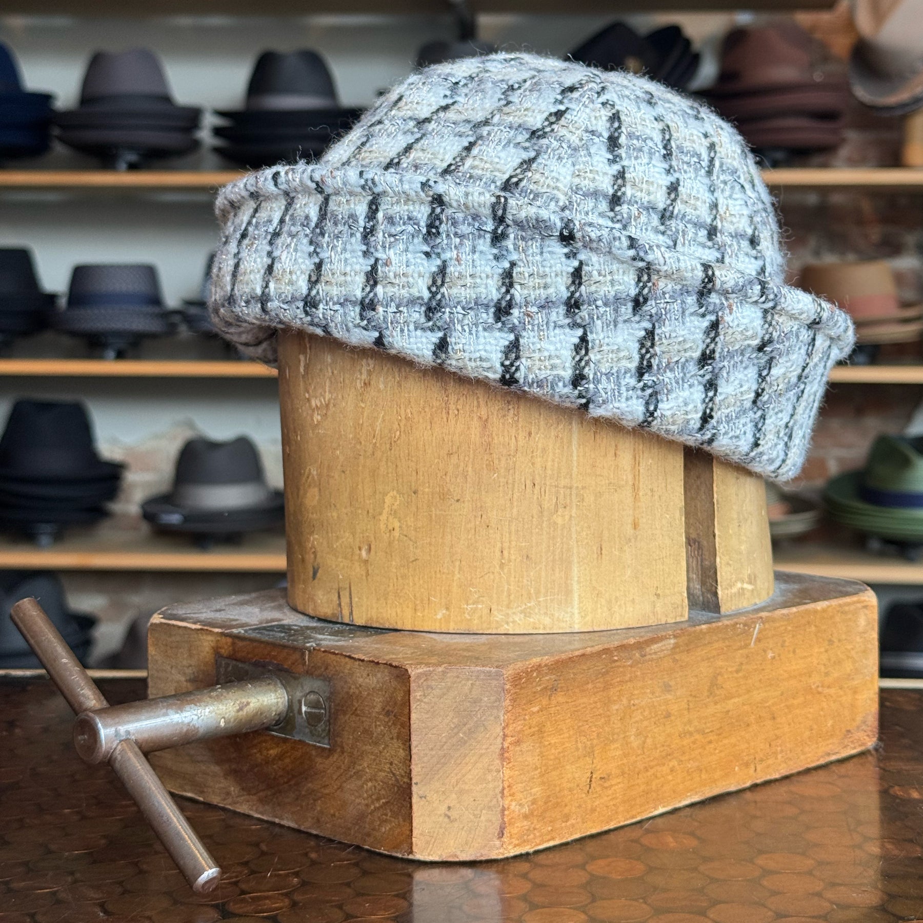 Tweed Sailor Cap displayed on a wooden hat block in a shop, featuring a textured gray and black patterned fabric, surrounded by various hats on shelves in the background.