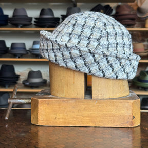 Tweed Sailor Cap in a black and white checkered pattern displayed on a wooden hat block with various hats blurred in the background.