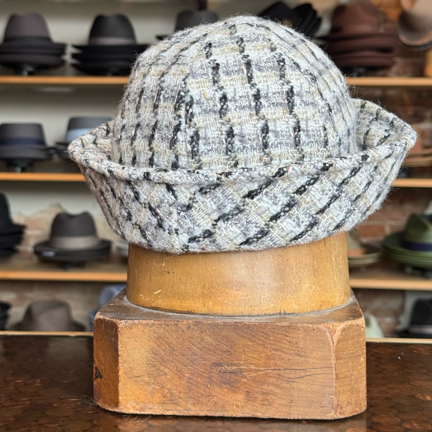 Tweed Sailor Cap made from textured gray and black tweed fabric with a rolled brim, displayed on a wooden hat stand in a shop with various hats on shelves in the background.