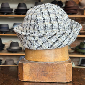 Tweed Sailor Cap made from textured gray and black tweed fabric with a rolled brim, displayed on a wooden hat stand in a shop with various hats on shelves in the background.