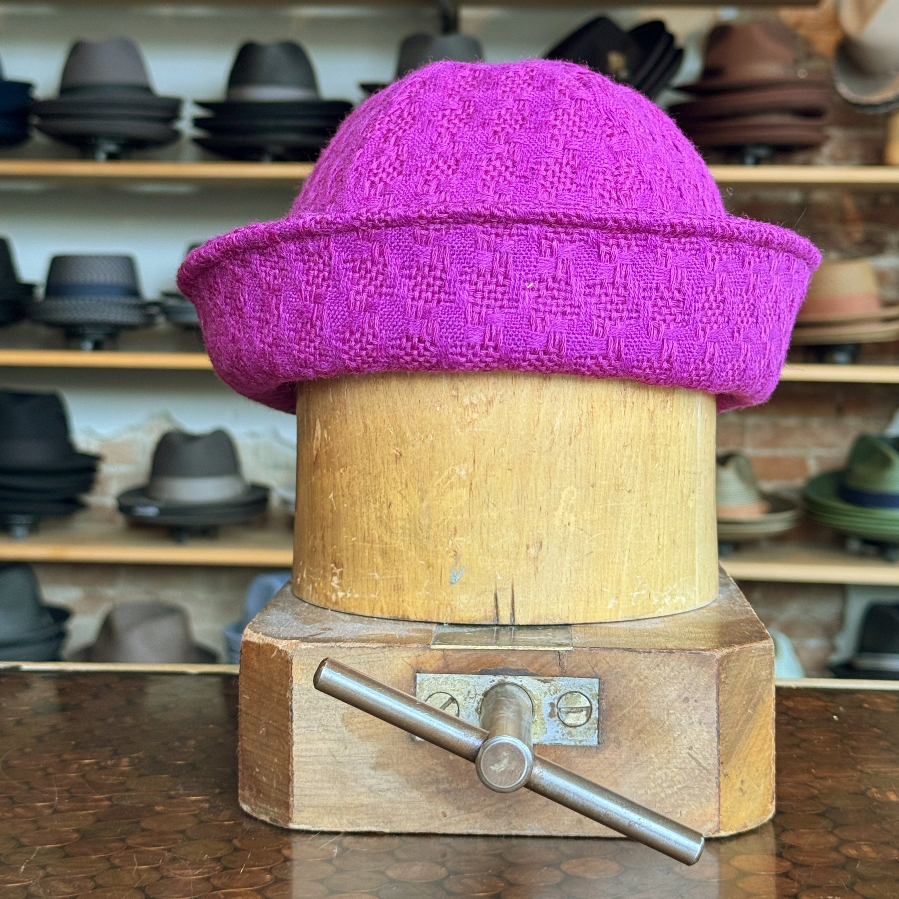 Tweed Sailor Cap in vibrant fuchsia pink displayed on a wooden hat block, showcasing textured woven fabric with a rolled brim, surrounded by a variety of hats in the background.