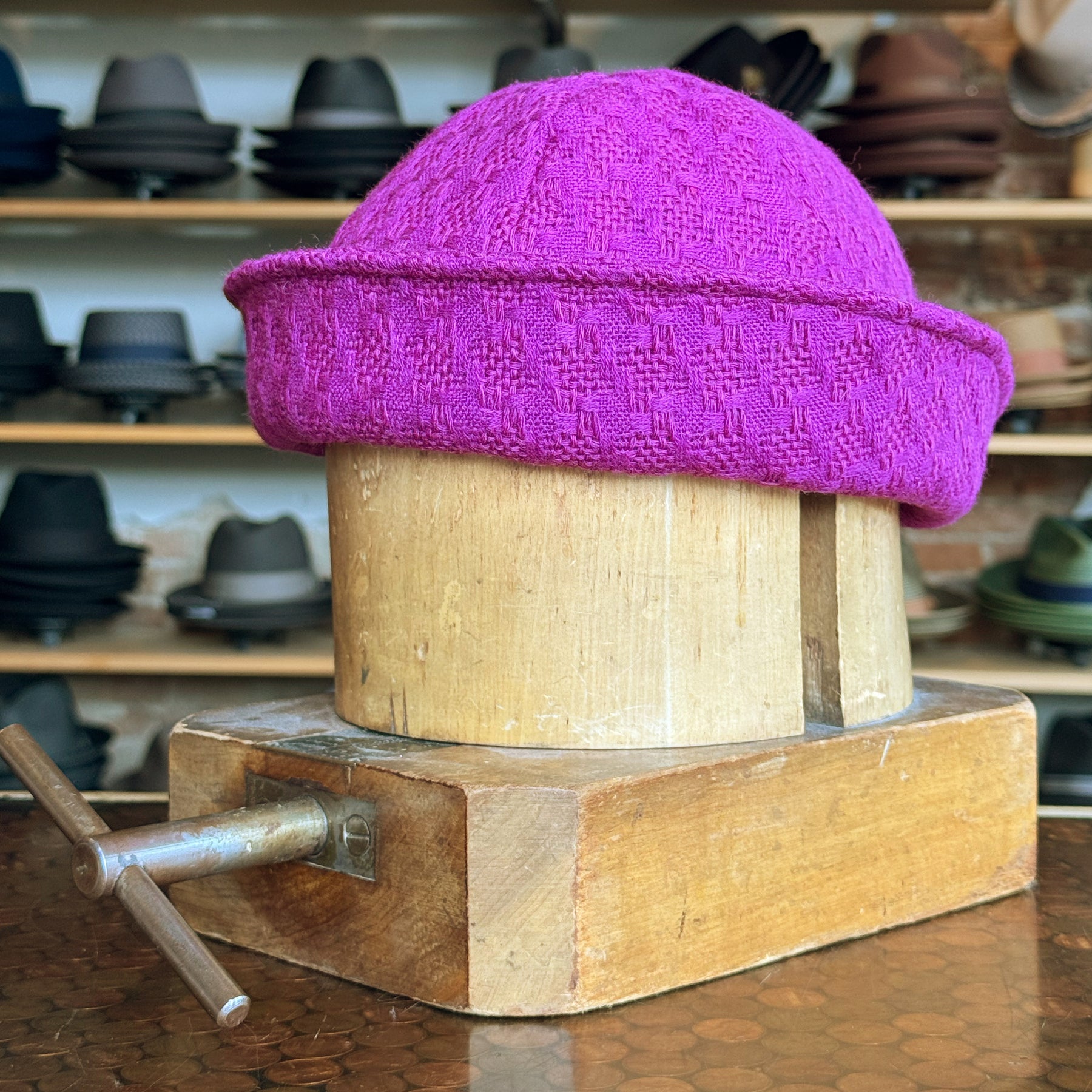 Tweed Sailor Cap in vibrant fuchsia displayed on a wooden hat block, showcasing its textured tweed fabric and rolled brim in a hat shop setting.