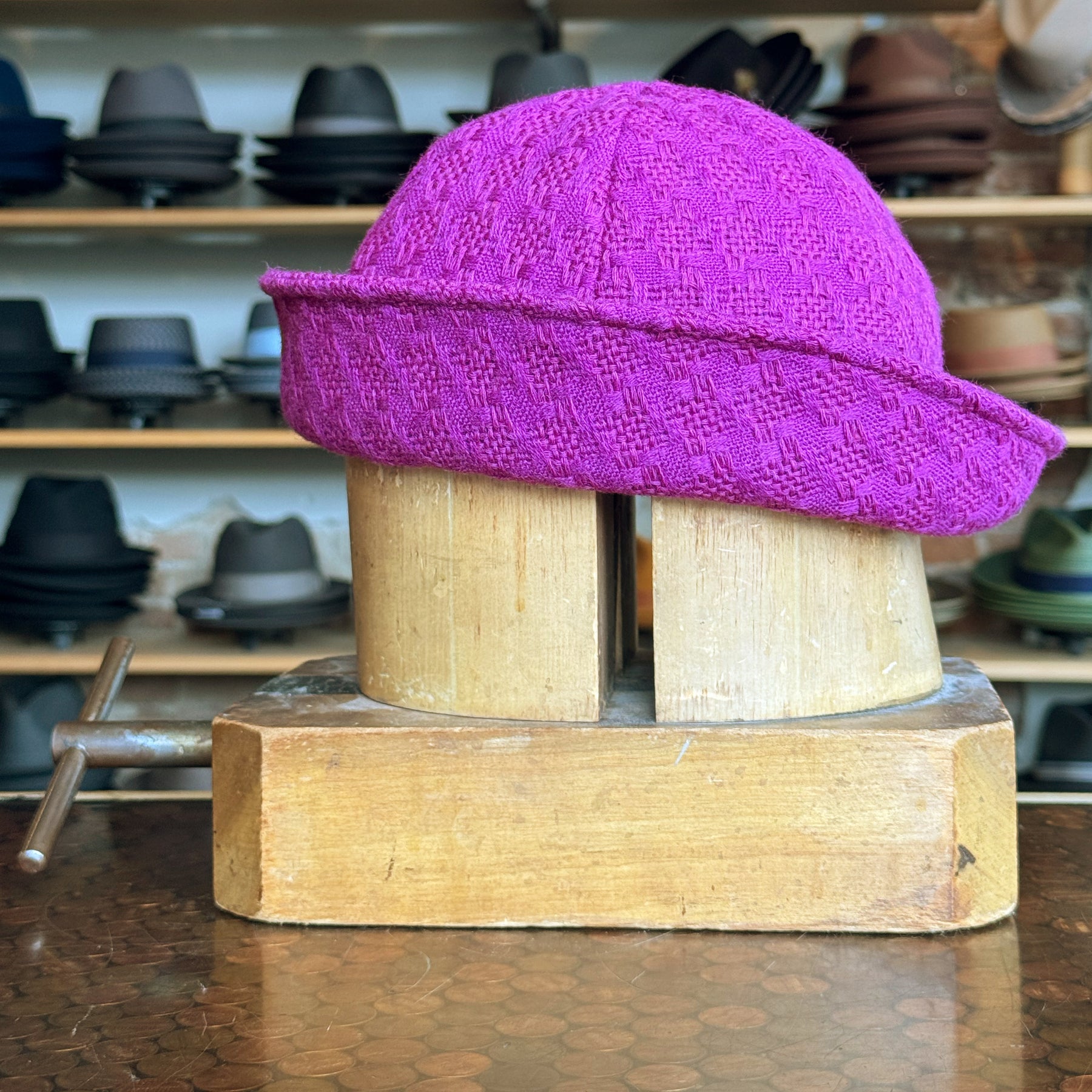 Tweed Sailor Cap in vibrant fuchsia with a textured pattern, displayed on a wooden hat block, surrounded by various hats in the background.