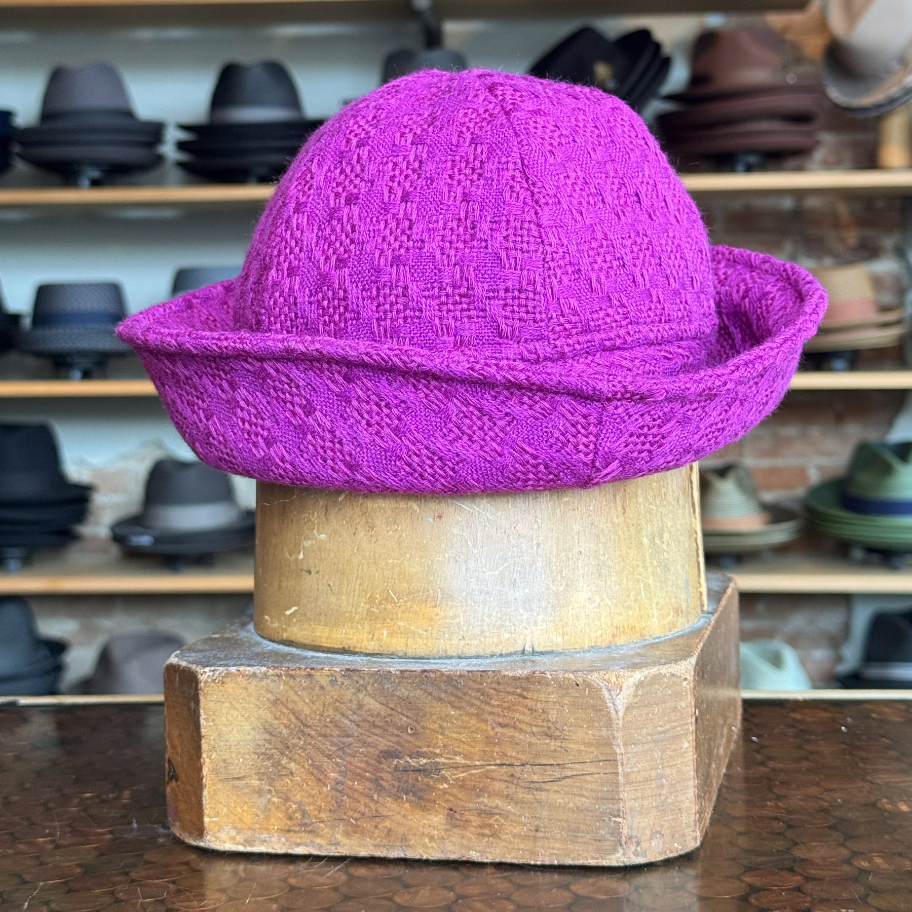 Tweed Sailor Cap in vibrant fuchsia with textured woven pattern, displayed on a wooden hat block, surrounded by various hats in the background.