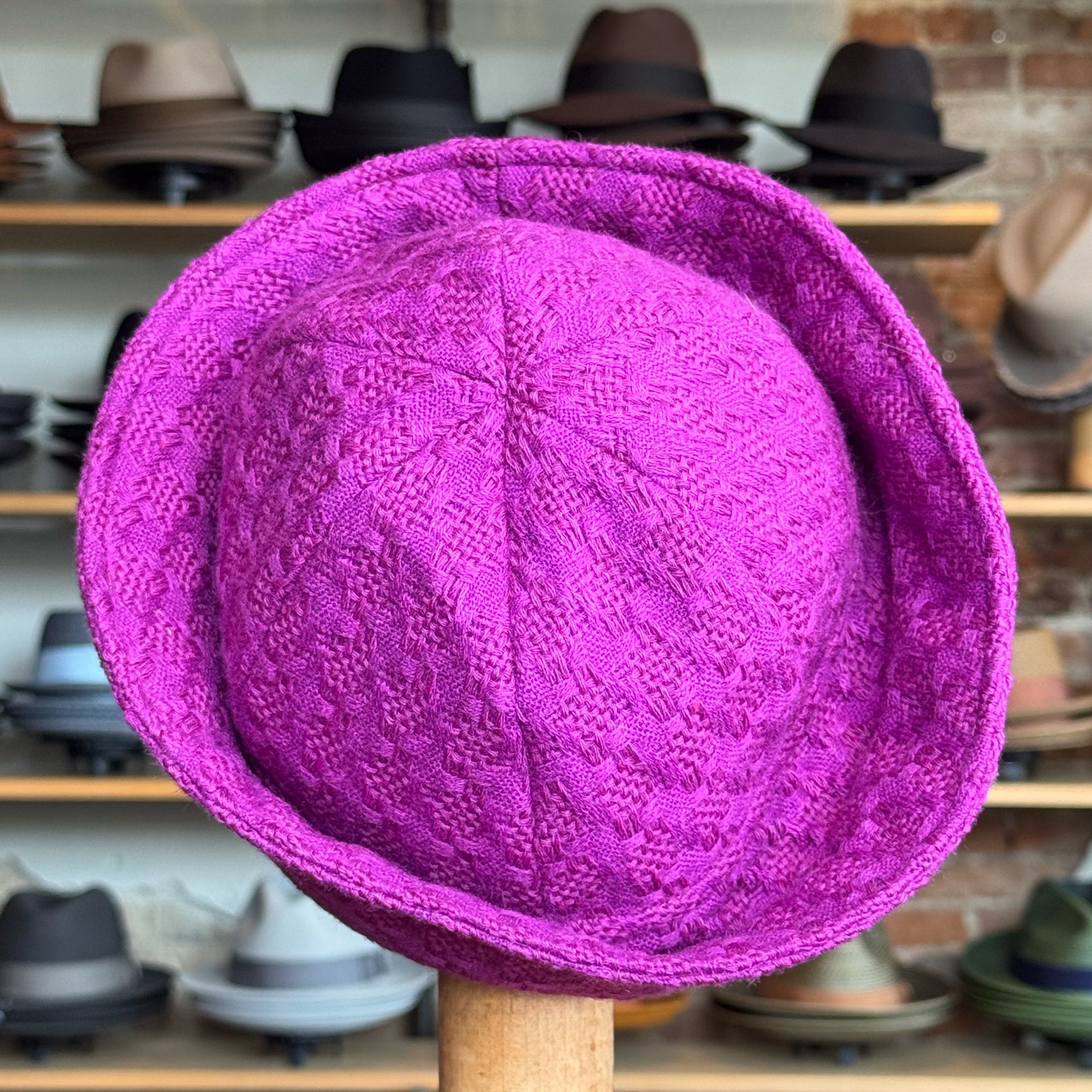 Tweed Sailor Cap in vibrant fuchsia color with textured weave pattern, displayed on a stand against a background of various hats on wooden shelves.