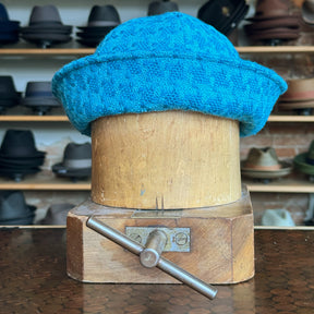 Tweed Sailor Cap in vibrant turquoise textured fabric displayed on a wooden hat block with a background of various hats on shelves in a store.