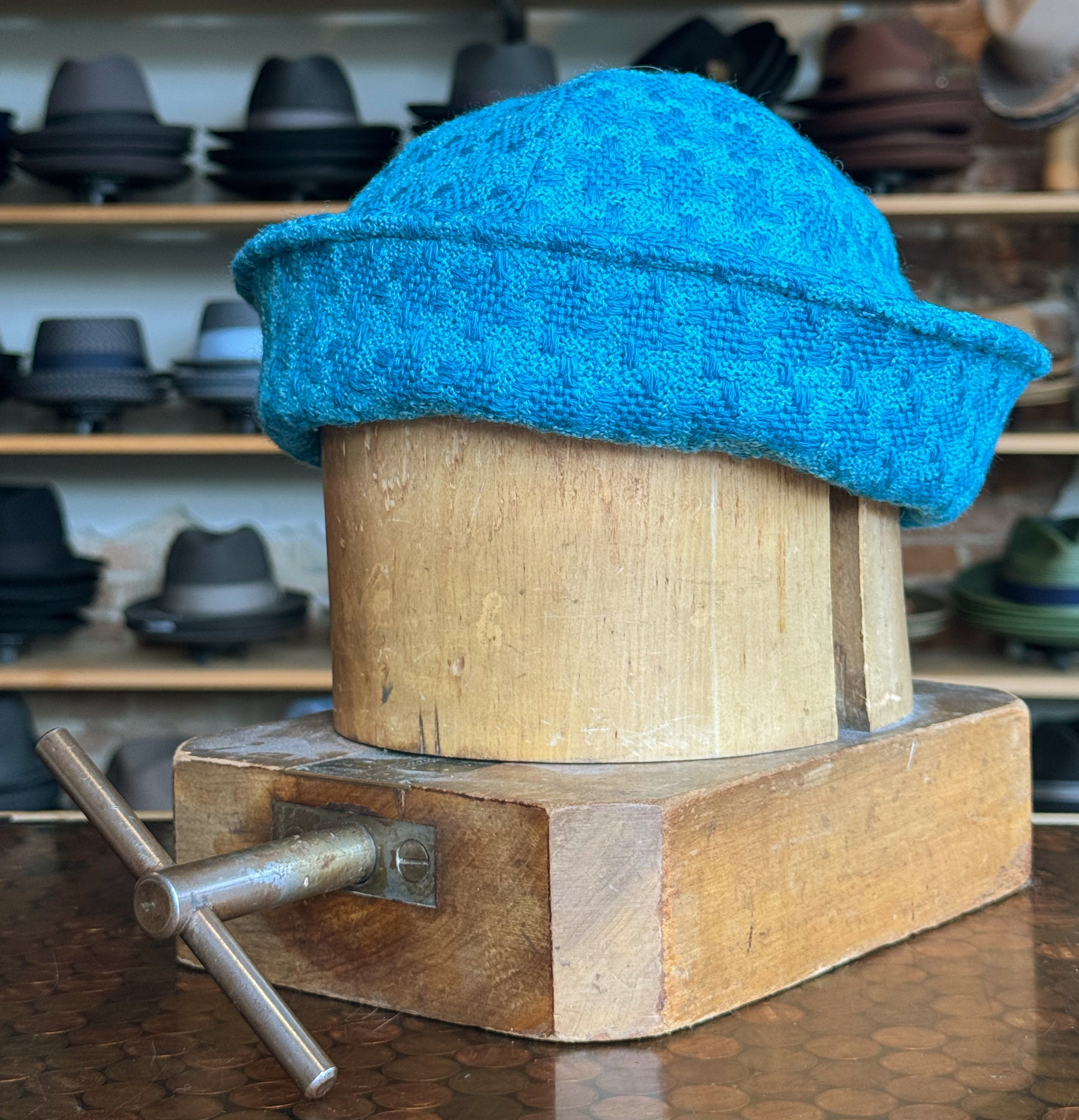 Tweed Sailor Cap in vibrant turquoise with a textured pattern, displayed on a wooden hat block in a shop filled with various brim hats and caps in the background.