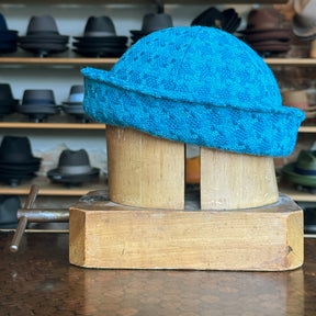 Tweed Sailor Cap in bright turquoise wool blend displayed on a wooden hat block, surrounded by various hats in the background on shelves.