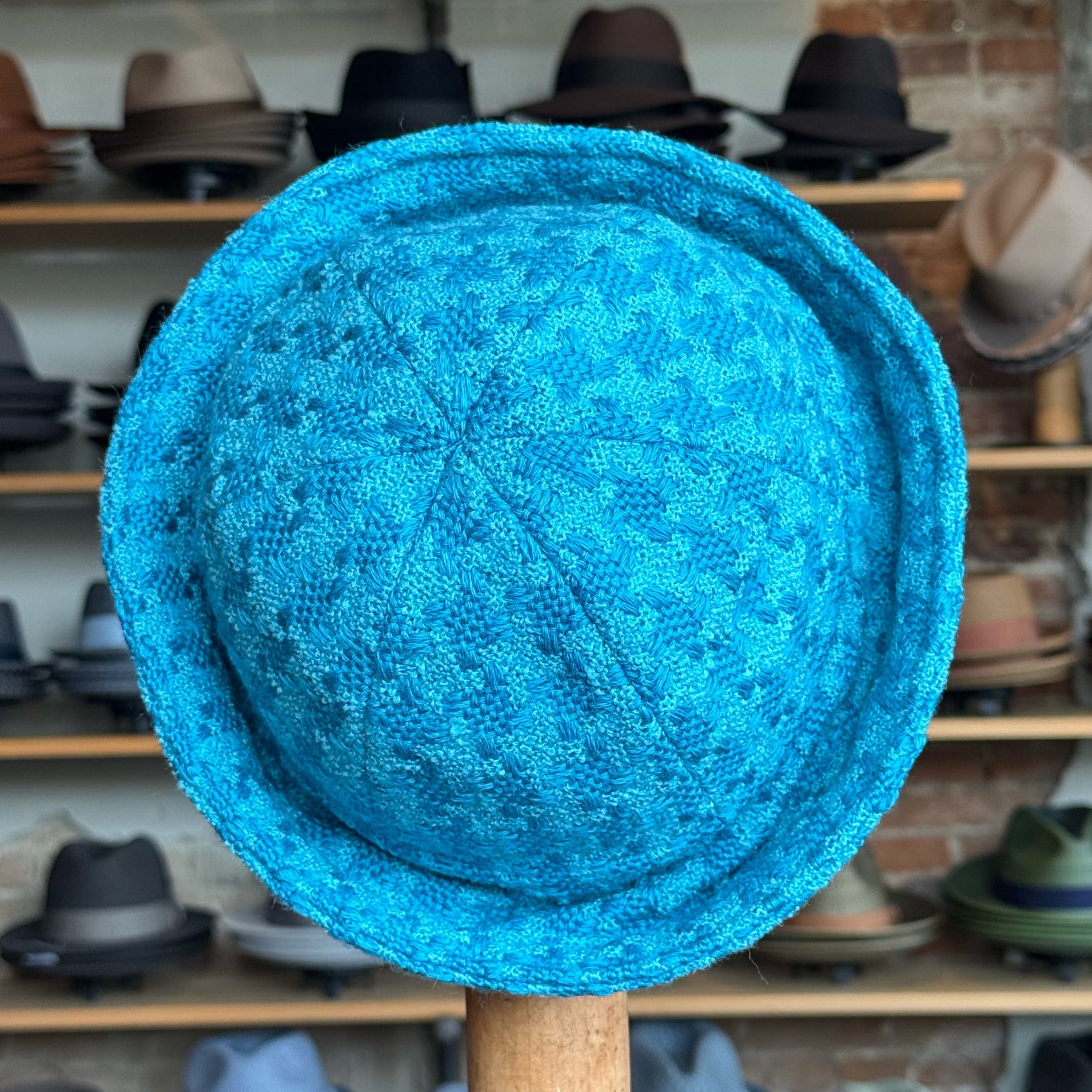 Tweed Sailor Cap in vibrant turquoise with textured woven fabric and a soft brim, displayed in a hat shop setting among various other hats and caps.