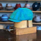 Vibrant Linen Hatteras flat cap in teal linen fabric displayed on a wooden hat block with shelves of various hats blurred in the background.