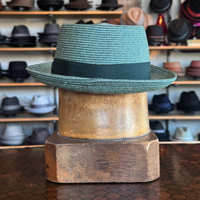 Waits Pork Pie hat in textured blue woven material with a black band, displayed on a wooden hat block with shelves of various hats in the background.
