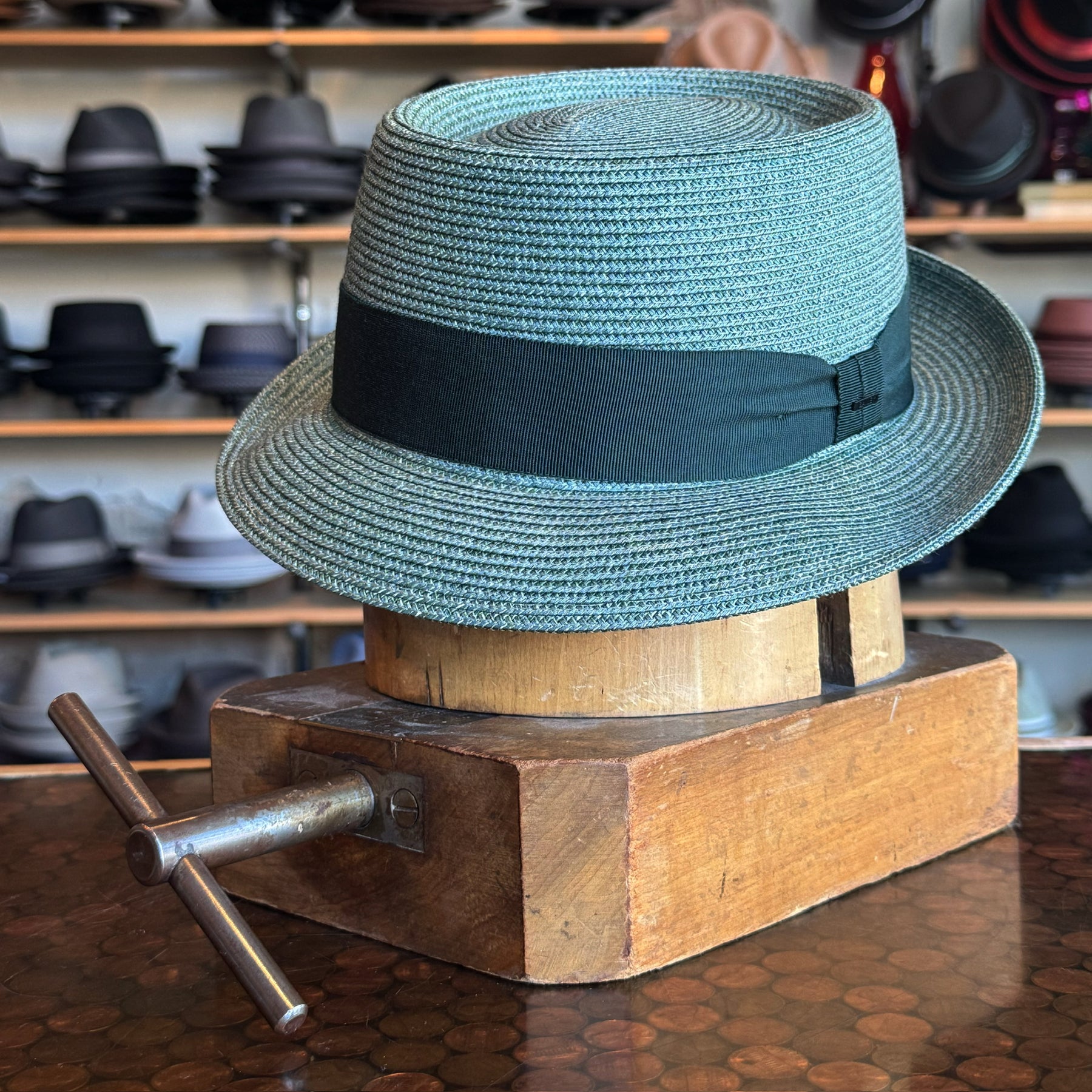 Waits Pork Pie hat in a light gray woven material with a black ribbon around the crown, displayed on a wooden hat block in a store with various hats on shelves in the background.
