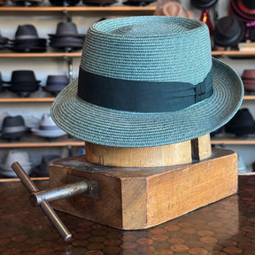 Waits Pork Pie hat in a light gray woven material with a black ribbon around the crown, displayed on a wooden hat block in a store with various hats on shelves in the background.