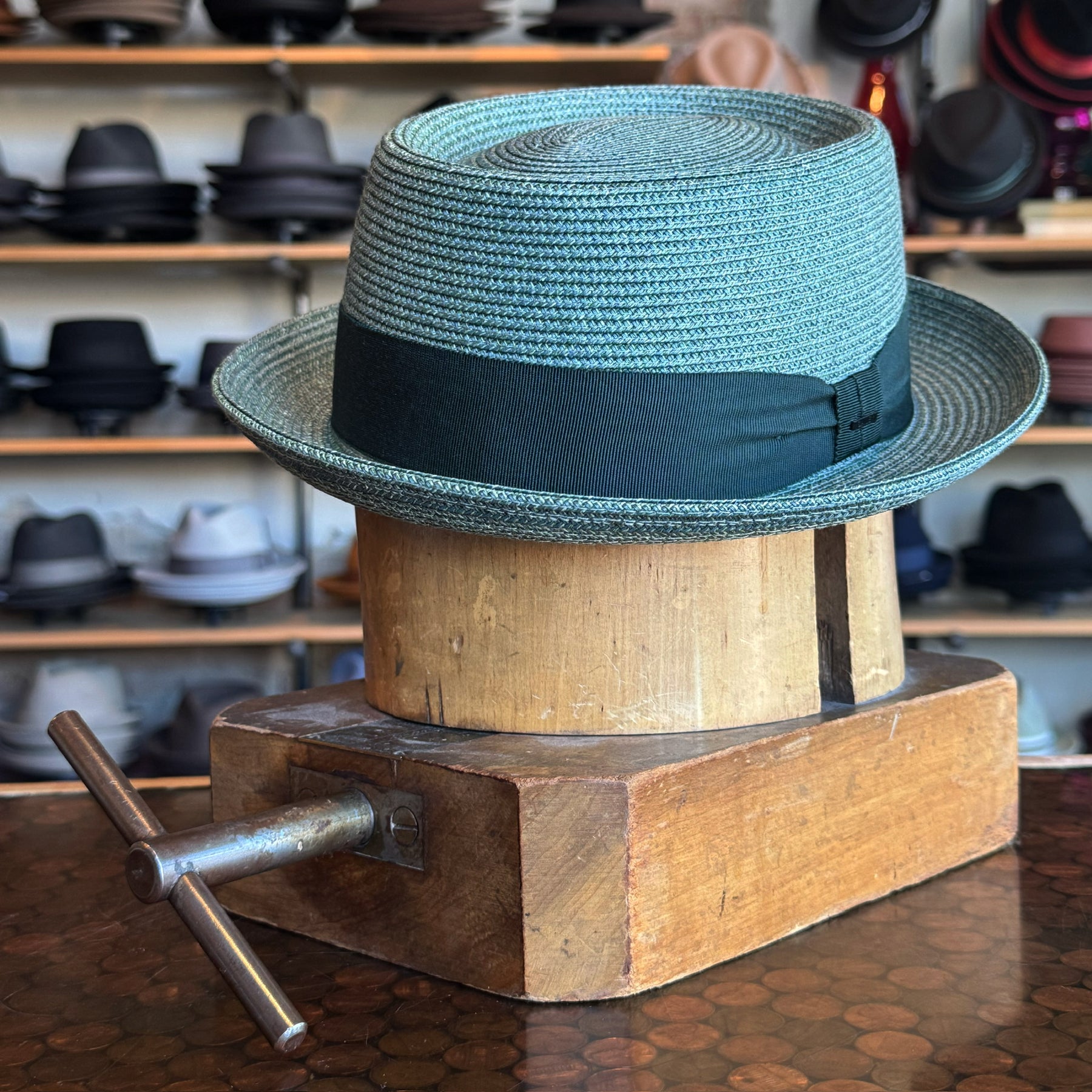 Waits Pork Pie hat in light blue with a black band displayed on a wooden hat block, surrounded by various hats on shelves in the background.