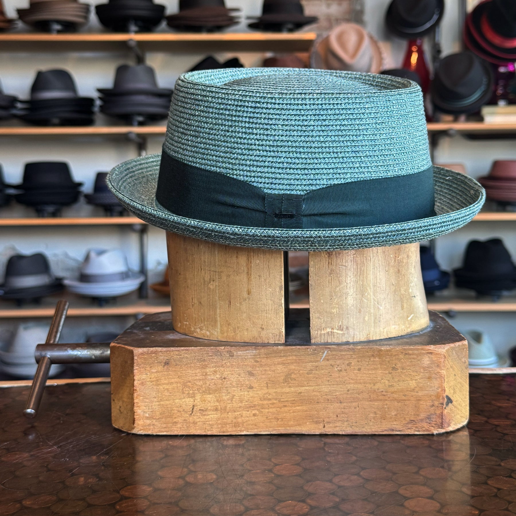 Waits Pork Pie hat in light teal woven material with a black ribbon band, displayed on a vintage wooden hat block in a shop filled with various hats in the background.