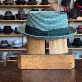 Waits Pork Pie hat in light teal woven material with a black ribbon band, displayed on a vintage wooden hat block in a shop filled with various hats in the background.