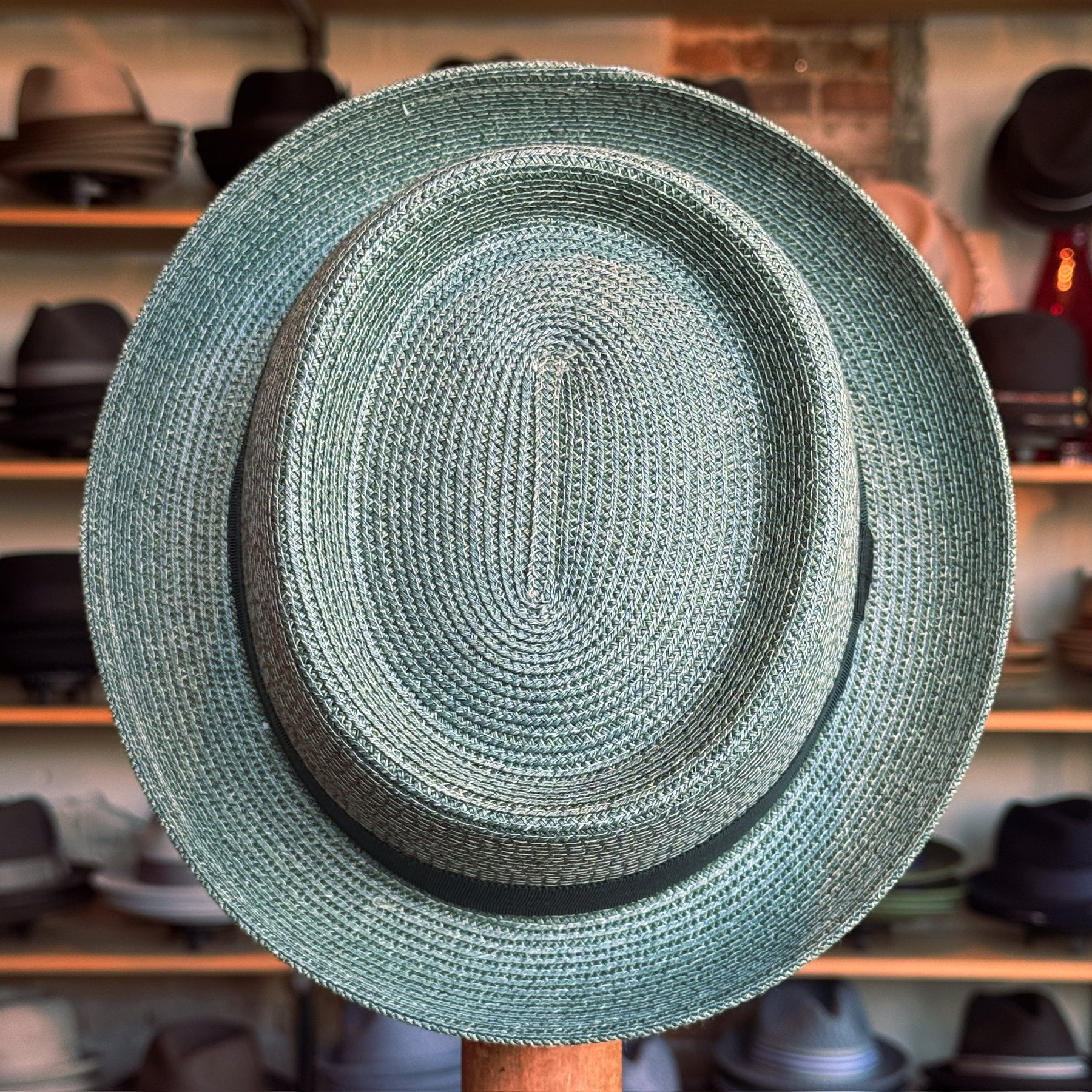 Waits Pork Pie hat in woven straw material with a classic black band, displayed in a shop setting with various other hats in the background.