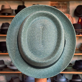 Waits Pork Pie hat in woven straw material with a classic black band, displayed in a shop setting with various other hats in the background.