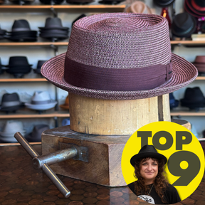 A stylish woven pork pie hat in shades of purple with a matching ribbon band, displayed on a wooden hat block in a shop with various other hats in the background, featuring the Waits Pork Pie.