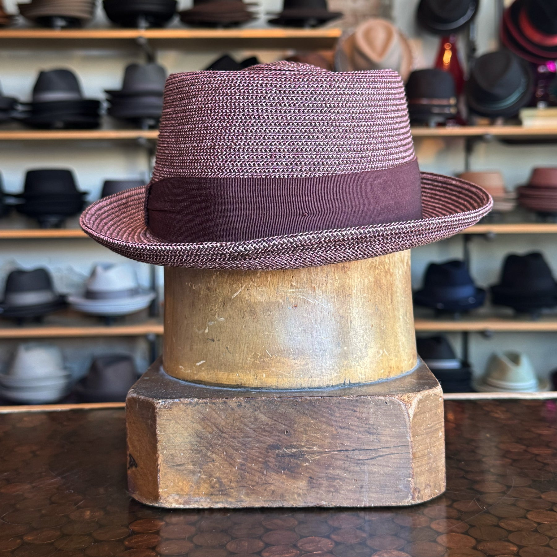 Waits Pork Pie hat in a textured maroon fabric with a wide matching band, displayed on a wooden hat block with shelves of various hats in the background.