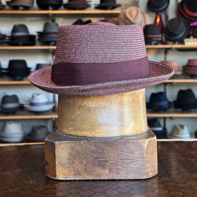 Waits Pork Pie hat in a textured maroon fabric with a wide matching band, displayed on a wooden hat block with shelves of various hats in the background.