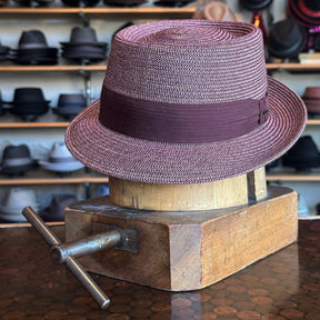 Waits Pork Pie hat in a woven burgundy and white pattern with a matching solid burgundy band, displayed on a wooden hat stretcher in front of shelves filled with various hats.