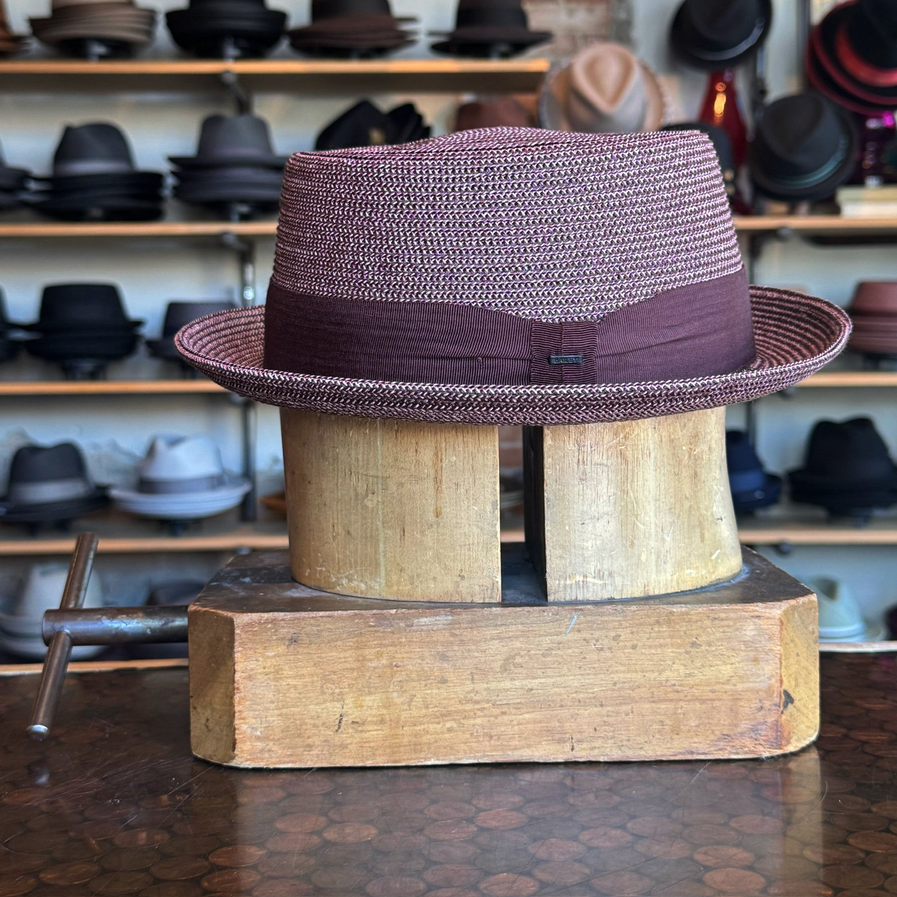 Waits Pork Pie hat in woven texture with a dark band, displayed on a wooden hat block in a shop filled with various styles of hats in the background.