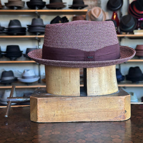 Waits Pork Pie hat in woven texture with a dark band, displayed on a wooden hat block in a shop filled with various styles of hats in the background.