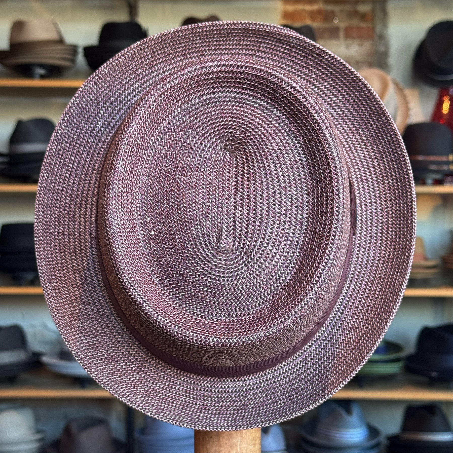 Waits Pork Pie hat crafted from woven material in a muted reddish tone, displayed in a hat shop with shelves of various hats in the background.
