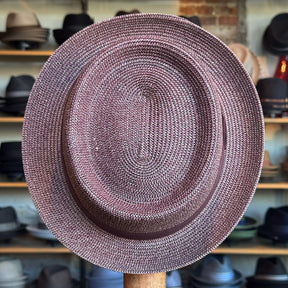 Waits Pork Pie hat crafted from woven material in a muted reddish tone, displayed in a hat shop with shelves of various hats in the background.