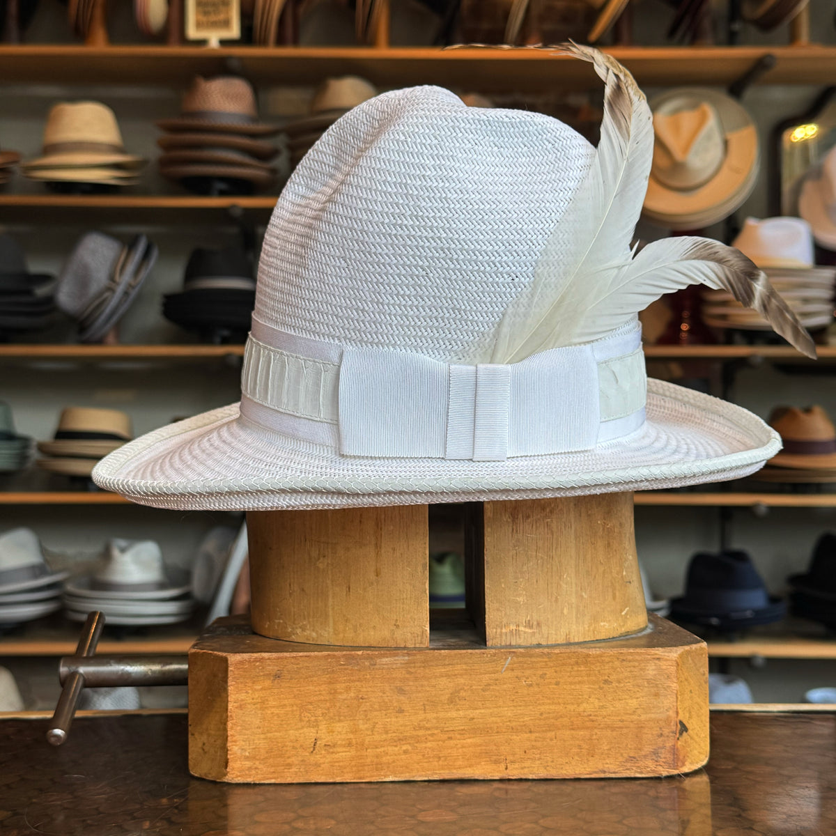 White on Rice Slouch Hat displayed on a wooden hat block, featuring a woven texture, wide brim, white ribbon band, and decorative white feathers in a stylish hat shop setting.