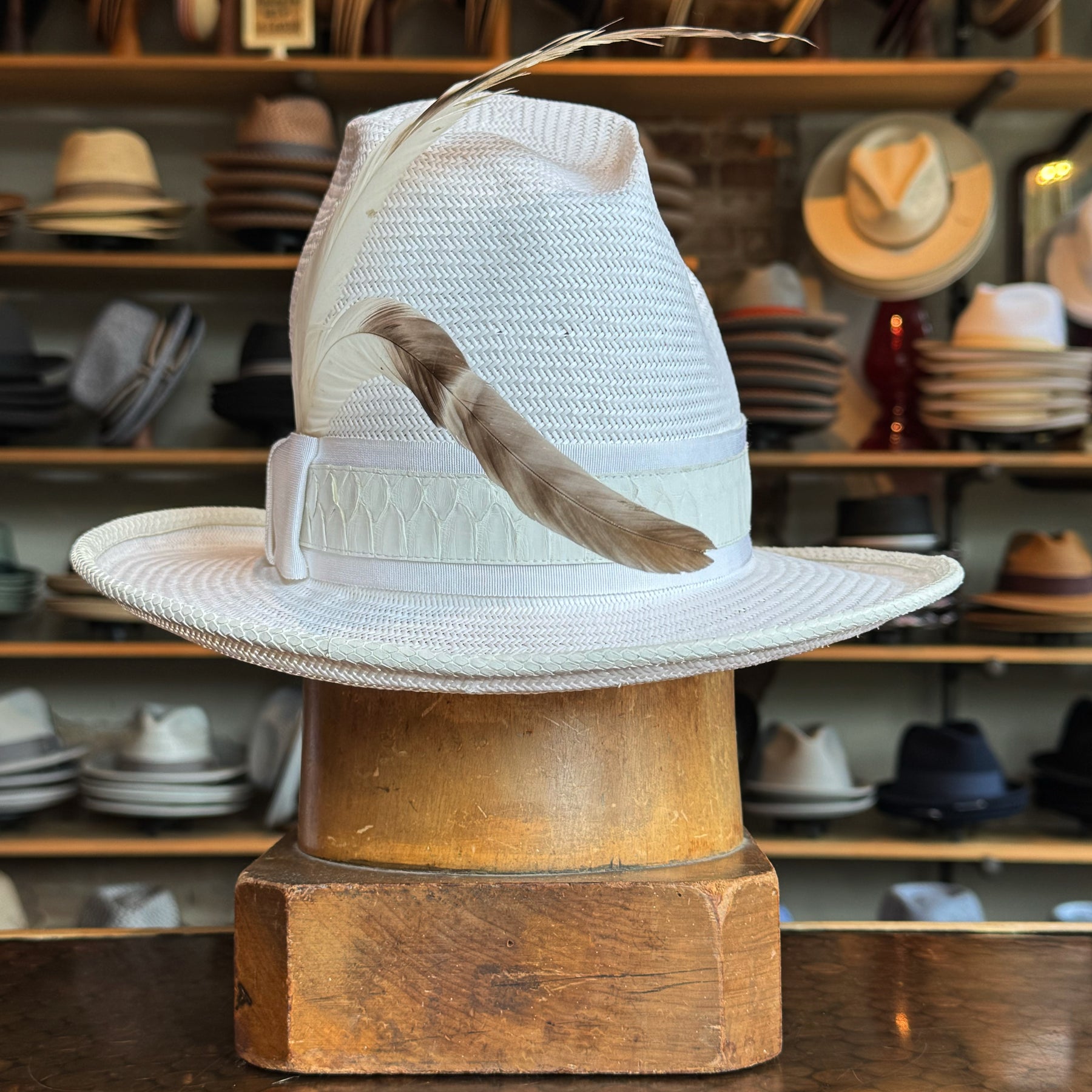 White on Rice Slouch Hat displayed on a wooden block with a decorative feather, surrounded by various hats in a store setting showcasing brim hats and accessories.