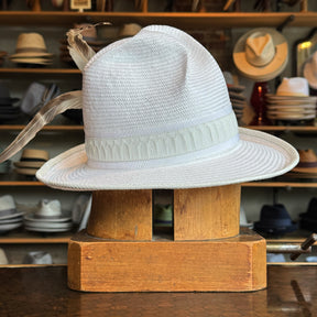 White on Rice Slouch Hat in a woven light material with a subtle feather embellishment and decorative band, displayed on a wooden stand in a hat shop with various hats in the background.