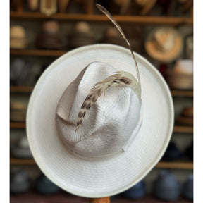 White on Rice Slouch Hat made of woven natural fibers with a wide brim and decorative feathers, displayed in a hat shop with various hats blurred in the background.