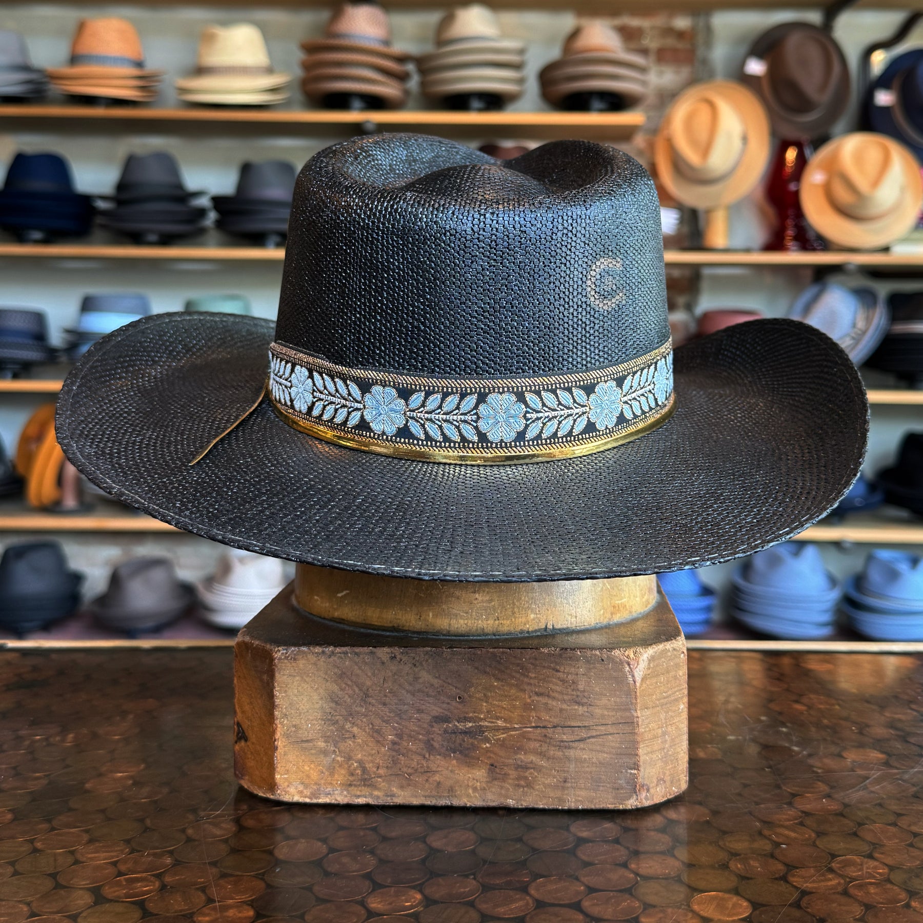 Wildflower black woven brim hat with blue floral patterned band displayed on a wooden stand in a hat store background.
