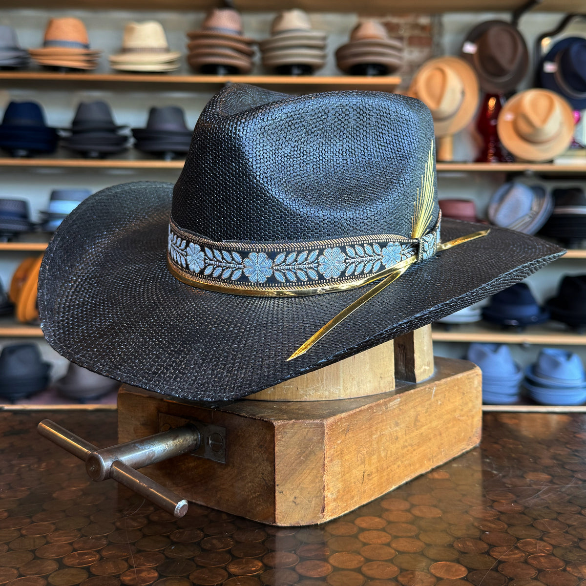 Black woven Wildflower hat with a decorative blue floral band and yellow feather, displayed on a wooden hat block with shelves of various hats in the background.
